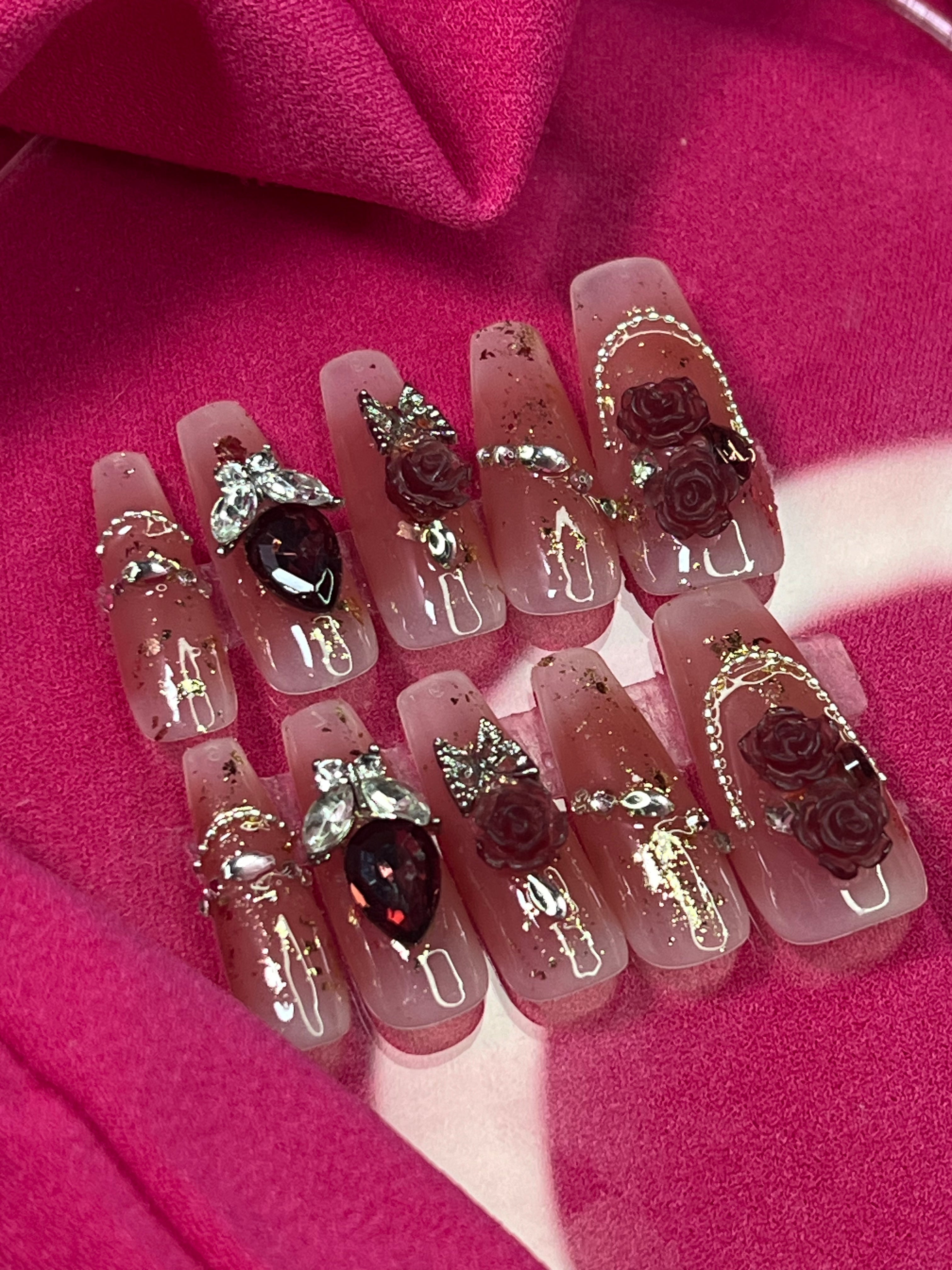 A set of Royal Palm Crush Press-on Nails featuring a coffin shape with pinkish tones, painted roses on the thumb and middle fingers, and a red heart-shaped gemstone on the ring finger, displayed on a vibrant pink background.
