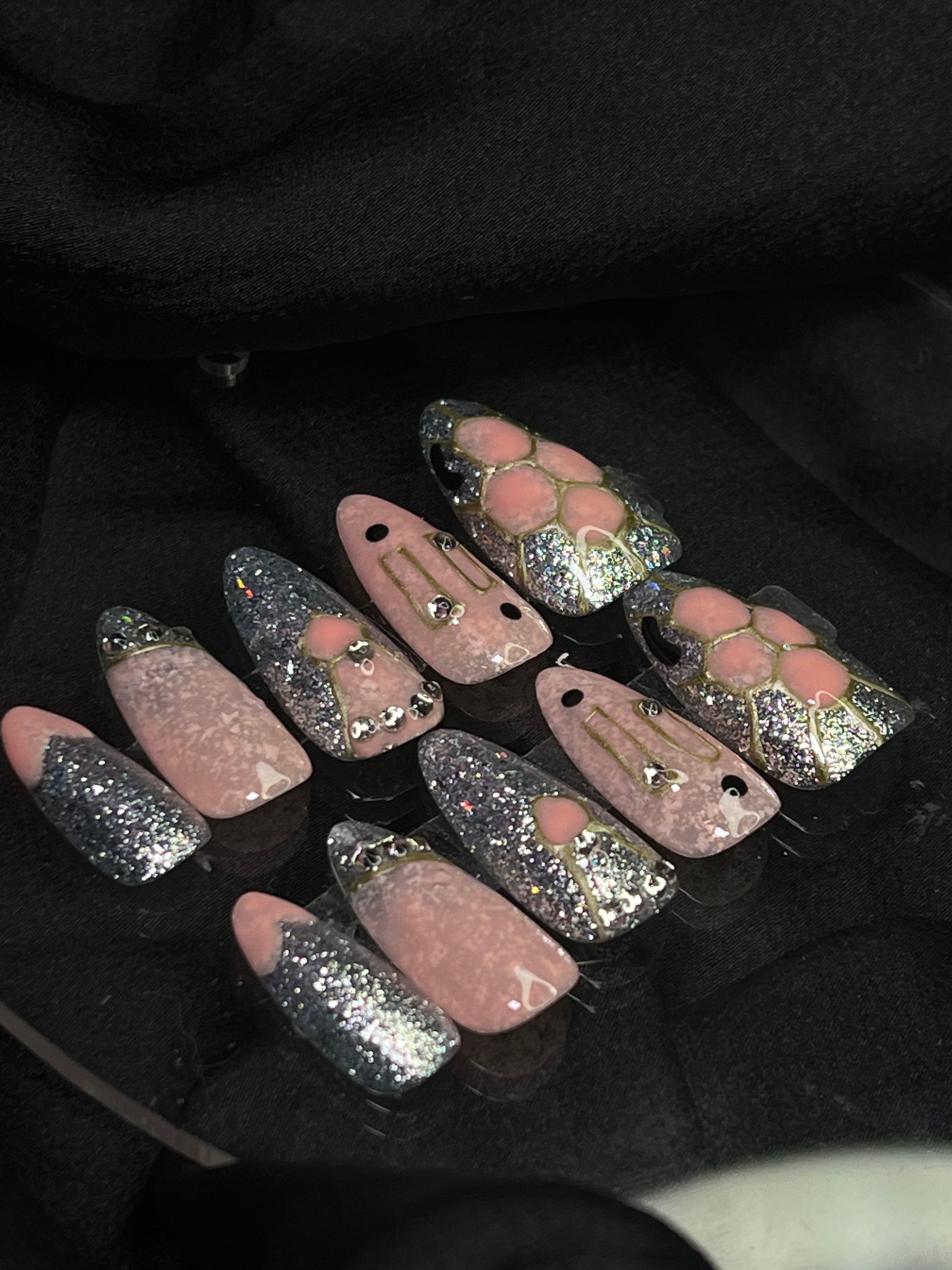 A set of Velvet Serpent Luxe Elegance Press-on Nails with a pinkish-red base, glitter accents, and serpent-like details, displayed on a vibrant pink fabric.